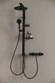 Schwarze Duscharmatur von Ideal Standard mit einer Handbrause, einem festen Duschkopf und Ablagen für Pflegeprodukte an der Wand montiert.